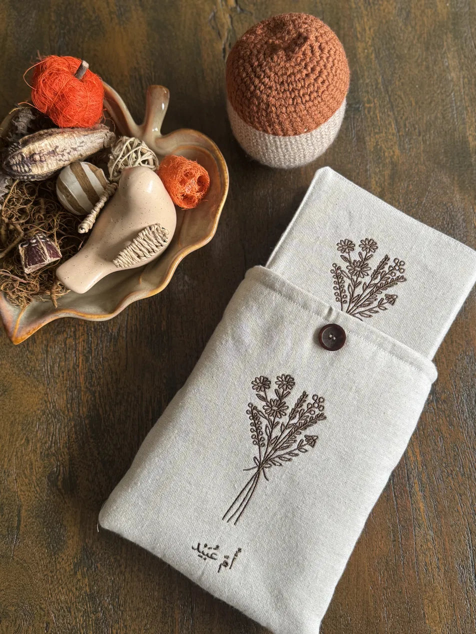A folded light beige linen fabric pouch or sleeve with a dark brown embroidered floral bouquet print and a small brown button closure, styled on a rustic wooden surface with decorative autumn props including a crocheted hat and a shell-shaped dish.